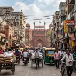 A bustling, crowded street in Old Delhi featuring green and yellow auto-rickshaws, pedestrians, and vendors, with the historic red sandstone walls of the Red Fort visible in the background under a hazy sky.